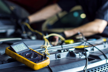 Closeup of yellow multimeter with cables on open hood. Mechanic is conducting diagnostics of vehicle at service station on background. Repairman is repairing car at auto repair shop workshop.