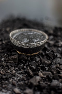 Close up of activated charcoal in a glass bowl on the wooden surface along with some raw powder of charcoal or coal spread on the surface.
