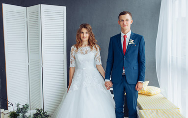 Fototapeta premium Beautiful bride in white dress and groom in suit, posing in white Studio interior, wedding