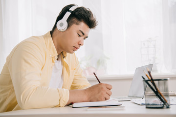 young, concentrated asian man in headphones writing in notebook white sitting at desk