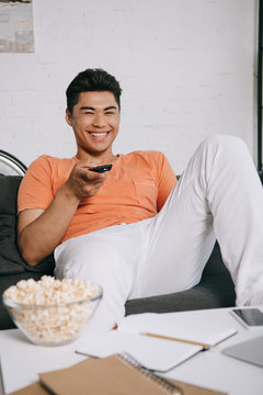 Cheerful Asian Man Watching Tv While Sitting On Couch Near Table With Bowl Of Popcorn