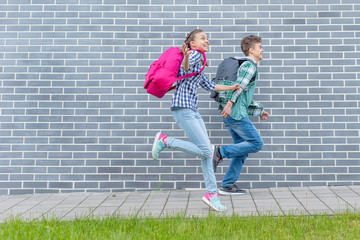 Two happy schoolchildren with backpacks run to school on street next to an Brick Wall. Cheerful cute children pupils Teen Girl and Boy Back to School. Concepts of friends, childhood and education. - 281060039