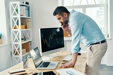 Middle age man in smart casual wear analyzing stock market data and talking on the phone while working in the office