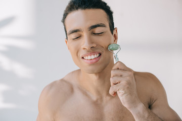 handsome mixed race man massaging face with jade roller and smiling with closed eyes