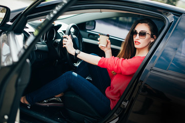 Fototapeta na wymiar Beautiful young woman in fashion sunglasses with coffee in hand sitting in black car. Female on road trip drinking coffee inside car. Lifestyle People Travel concepts