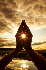 Tin namaste close-up, the sun shines between the fingers with stunning rays. Silhouette against the setting sun, sky and river. Concept of amateur yoga, healthy lifestyle, meditation practice.  - obrazy, fototapety, plakaty
