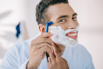 portrait shot of handsome man in bathrobe shaving face with razor while smiling and looking at...
