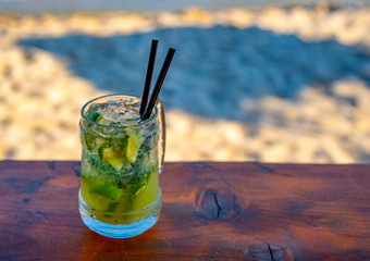 A glass with a refreshing mojito cocktail on the bar counter on the background of a sandy beach.