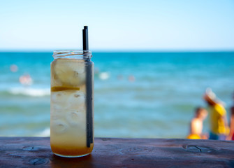 A jar of lemonade, in which float lemon and ice, with two black tubes, on the bar against the sea.