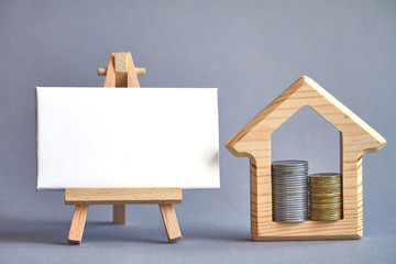 Wooden figure of house with two columns of coins inside and white board on miniature easel on gray background, the concept of buying or renting a building, selective focus