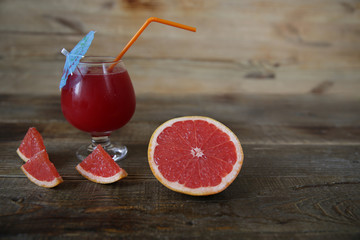 a glass of fresh red grapefruit juice with a straw and an umbrella and sliced ​​half a grapefruit on a rustic wooden background. With copy space for text. Healthy food, diet concept, detox