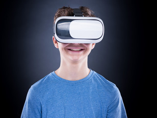 Happy teen boy wearing virtual reality goggles watching movies or playing video games, on black background. Cheerful teenager looking in VR glasses. Funny child experiencing 3D gadget technology.