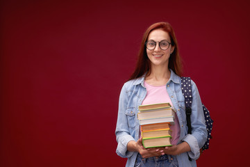 portrait of a beautiful girl student with a backpack and a stack of books in his hands is smiling at the red background.