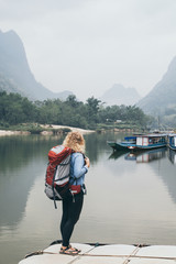 Caucasian blonde woman with backpack stands on boat pier in Muang Ngoi village, Laos