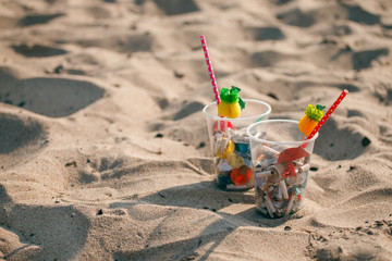 Plastic cups with straw and tropical decor and Garbage on the beach, trash, plastic, bottle, foam, rubbish, sigarette stub. Ecological creative concept, trash cocktail on the beach
