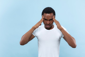 Half-length close up portrait of young african-american man in white shirt on blue background. Human emotions, facial expression, ad concept. Suffering from headache, heavy thoughts, mental problems.