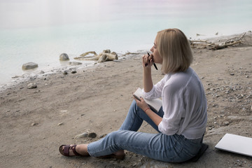 Beautiful blonde girl sits on the shore of the lake and writes in a notebook.