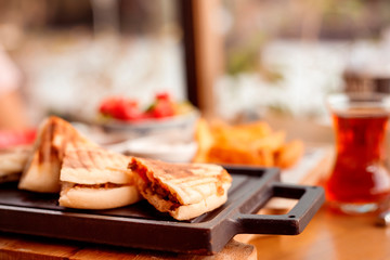 quesadilla sandwich with meat lies on black board on table in outdoor cafe. Turkish tea in glass, potatoes and vegetable salad next to it