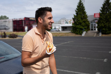 Handsome young brunette man eating hot dog in the parking lot near the gas station.