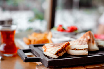 quesadilla sandwich with meat lies on black board on table in outdoor cafe. Turkish tea in glass, potatoes and vegetable salad next to it