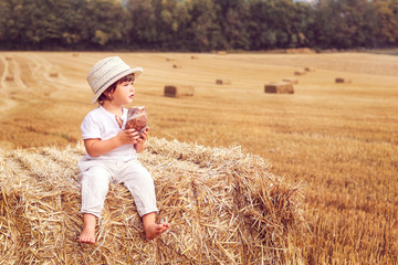 Cute little boy in straw hat eating bread sitting on hay stack in harvested yellow wheat field. Summer lifestyle. Child nutrition. Rural scene. Copy space