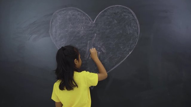 Back view of female elementary school student drawing a heart symbol on the chalkboard in the classroom at school. Shot in 4k resolution