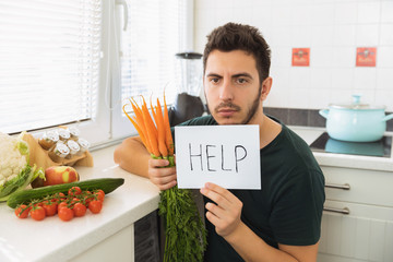 A young handsome man sits in the kitchen with a sad face and asks for help. The guy does not want to go on a healthy lifestyle and eat fresh vegetables.