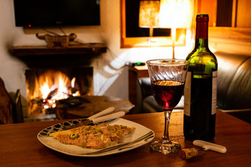 Foreground with bottle and glass of wine and pizza. Defocused background with fireplace with high heat.
