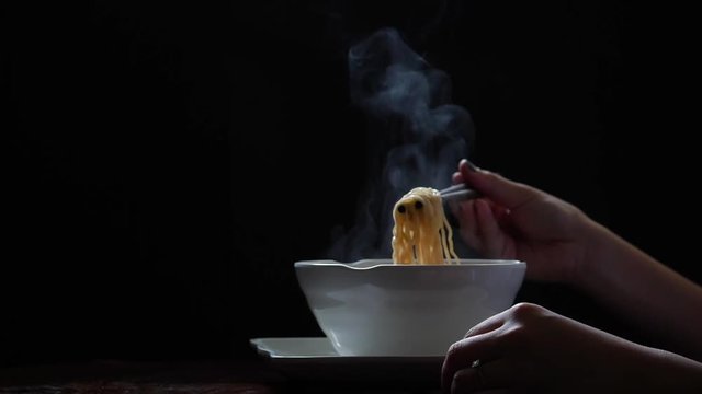 Asian girl is eating noodles with chopsticks, steam and smoke in white bowl on black background, junk food concept, Shooting at a rate of 180 fps.