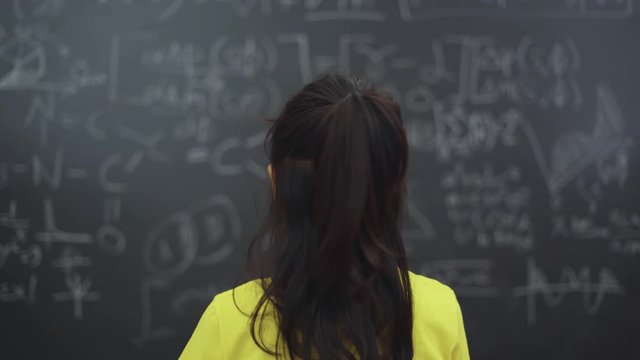 Back view of thoughtful female elementary school student trying to solve question math on the chalkboard in the classroom at school. Shot in 4k resolution