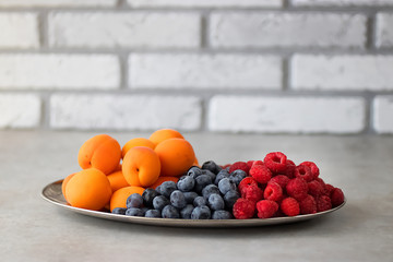 Fresh apricots, raspberries and blueberries on an oval metal plate. Gray background.