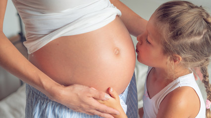 Authentic shot of a lovely little daughter is touching and kissing mother's pregnant belly, she is feeling baby at mother tummy and awaiting the birth of her little brother or sister.