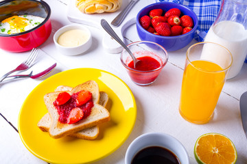 Café da manhã com ovos fritos, pão e manteiga, morangos, leite, cereais, torrada com geléia de morango e suco de laranja sobre a mesa de madeira branca