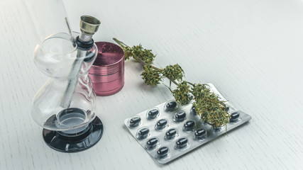 Marijuana buds, bong, grinder and pills lie on the white table.