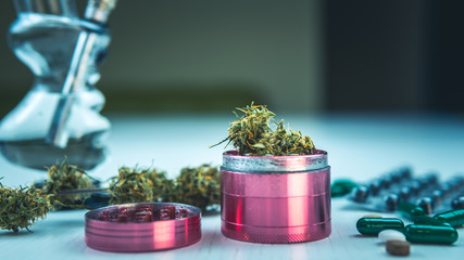 Marijuana buds, bong, grinder and pills lie on the white table.