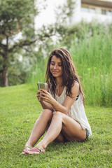 Fototapeta premium Portrait of cheerful brunette girl sitting on the garss in summer park. Close-up photo of young beautiful woman chatting and listening to music on her smartphone outdoors.
