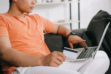 cropped view of young man using laptop and holding credit card while sitting on sofa at home