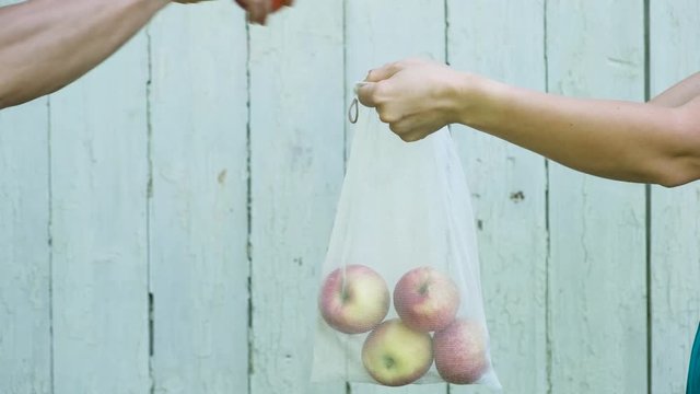 Woman's hand holds reusable recycled mesh produce bag, and man's hand puts apples in it on wooden background.