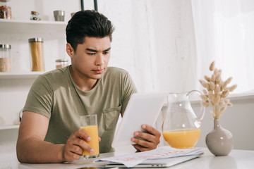 handsome asian man looking at digital tablet while sitting at kitchen table and holding glass of orange juice