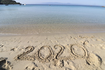 Happy New Year 2020, lettering on the beach with wave and clear blue sea. Numbers 2020 year on the sea shore, message handwritten in the golden sand on beautiful beach background. New Years concept.
