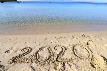 Happy New Year 2020, lettering on the beach with wave and clear blue sea. Numbers 2020 year on the sea shore, message handwritten in the golden sand on beautiful beach background. New Years concept.