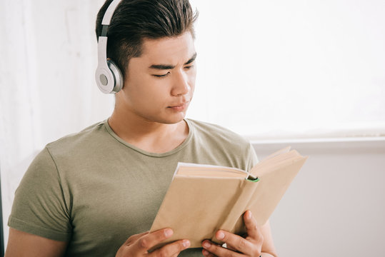 young asian man reading book while listening music in headphones