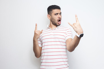 Fototapeta premium Portrait of funny handsome bearded young man in striped t-shirt standing with rock and roll horns gesture and looking at camaera with surprised face. indoor studio shot, isolated on white background.