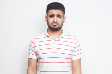 Portrait of sad updet or bored bearded young man in striped t-shirt standing and looking at camera with dissatisfied sadness face. indoor studio shot, isolated on white background.