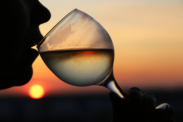 Silhouette of woman with misted glass of white wine at sunset, colorful sky and sun reflected in the drink. Girl drinking alcohol, romantic dinner at the resort, luxury life