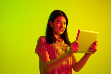 Beautiful female half-length portrait isolated on yellow studio background in neon light. Young emotional woman. Human emotions, facial expression concept. Using tablet for talking or internet serfing