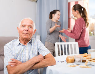 Upset senior man sitting, woman with mother quarreling on background
