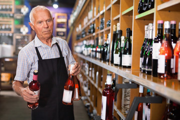 Confident elderly salesman of wine house arranging wine bottles on shelves rack