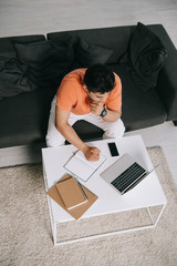 overhead view of young man writing in notebook and using laptop while sitting on sofa near desk