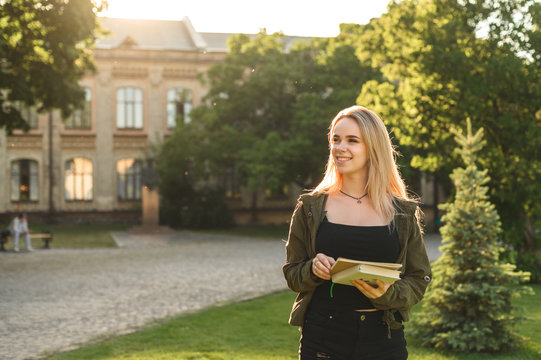 Beautiful female student with broad smile holding notebooks and looking aside in the college garden. Smiling attractive cute student girl with books and textbooks in the park near the university.
